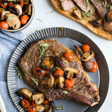 Steak, mushrooms, tomatoes and herbs on a tin plate with a bowl of mushrooms and tomatoes next to it and sliced meat on a cutting board, on top of a white background