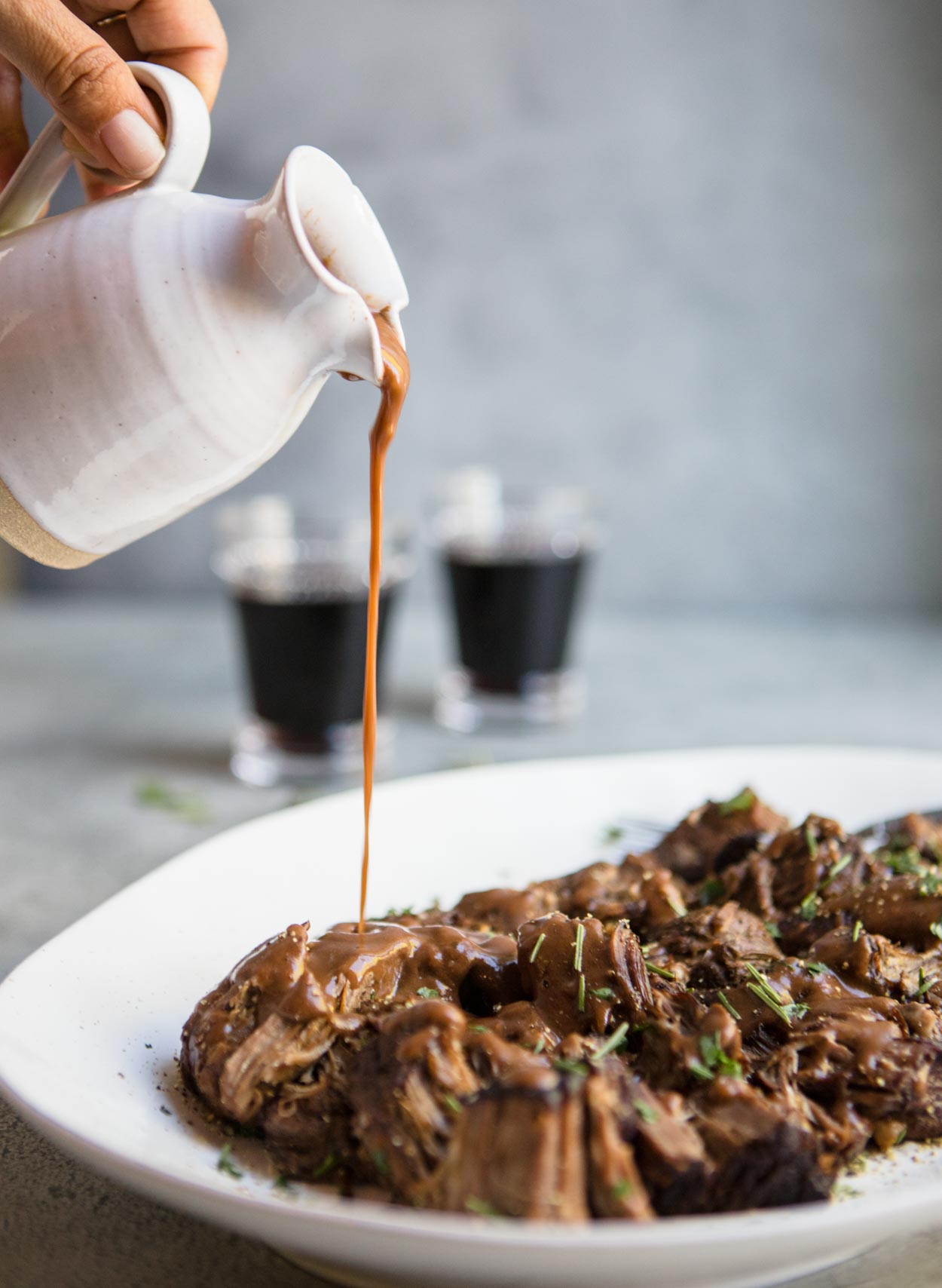 Gravy pouring over pot roast on a white platter set against a gray background and 2 wine glasses int the background