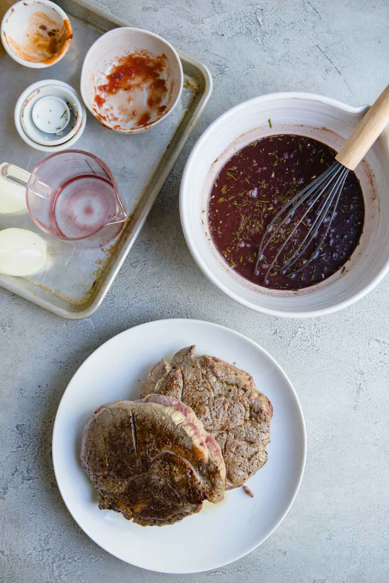 Seared pot roast on a white plate and a sauce mix in a white bowl set against a gray background