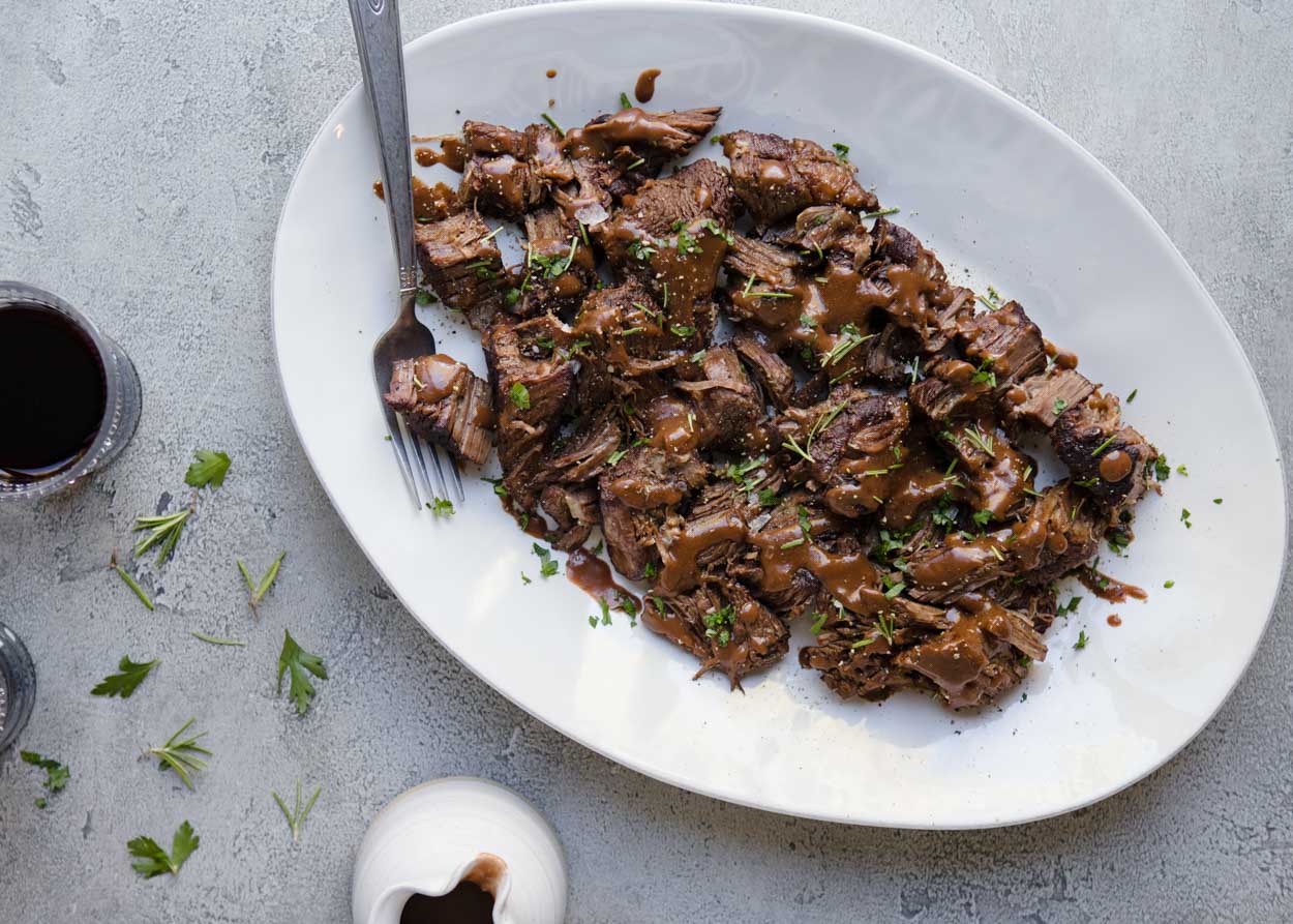 Pot roast with gravy on a white platter with fork set against a gray background