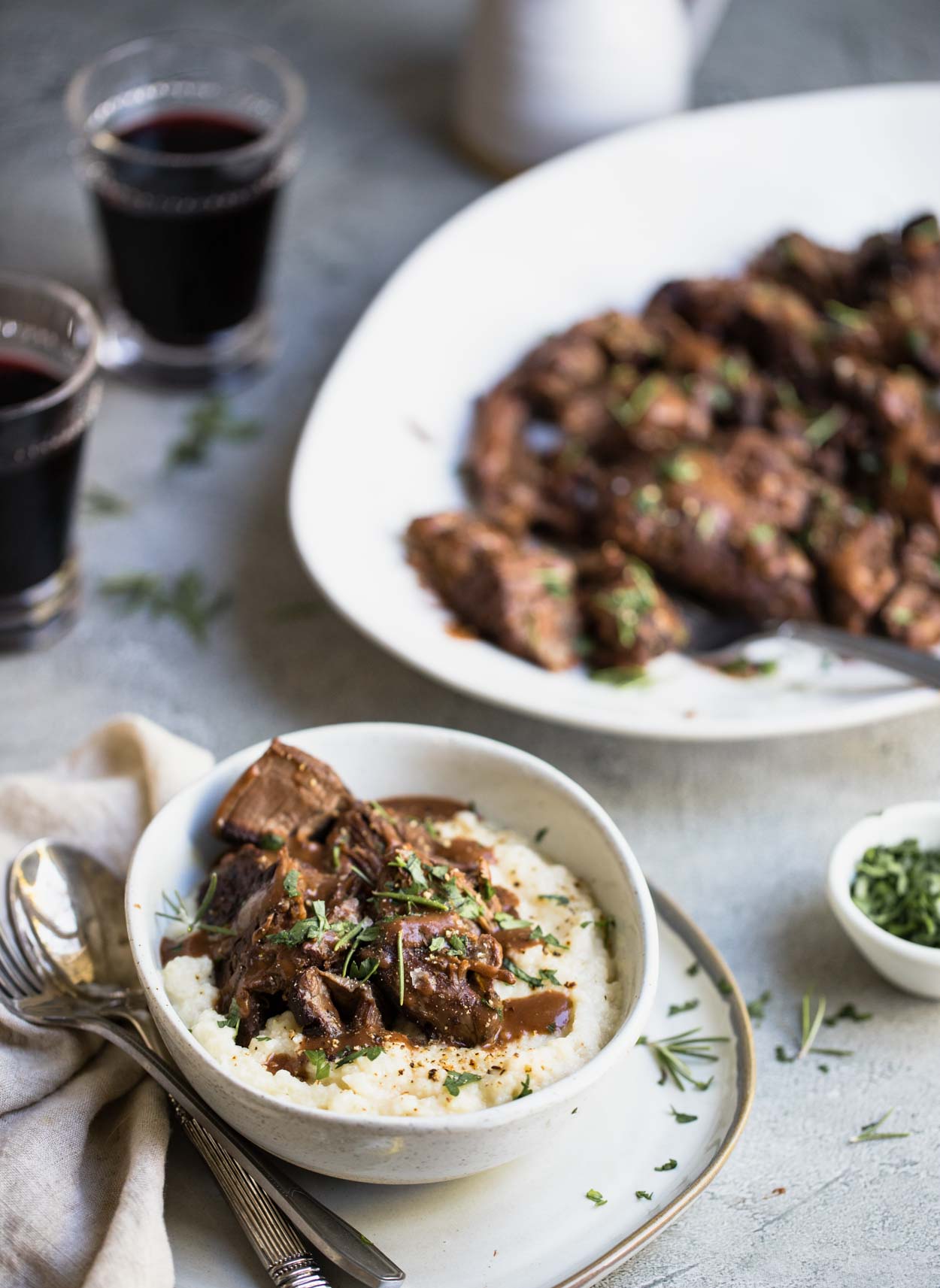 Pot roast and mashed potatoes with gravy in a white bowl and pot roast on a white platter with 2 wine glasses in the background