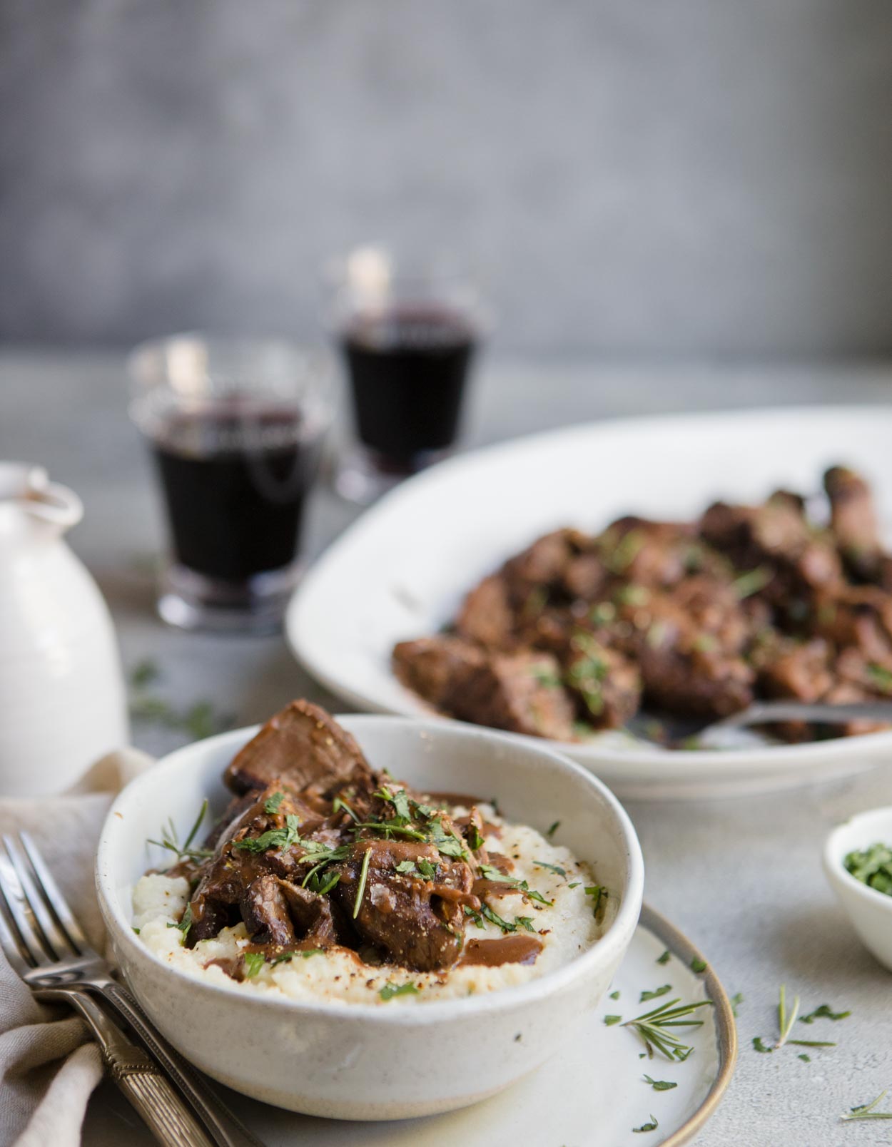 Pot roast and mashed potatoes with gravy in a white bowl and pot roast on a white platter with 2 wine glasses in the background