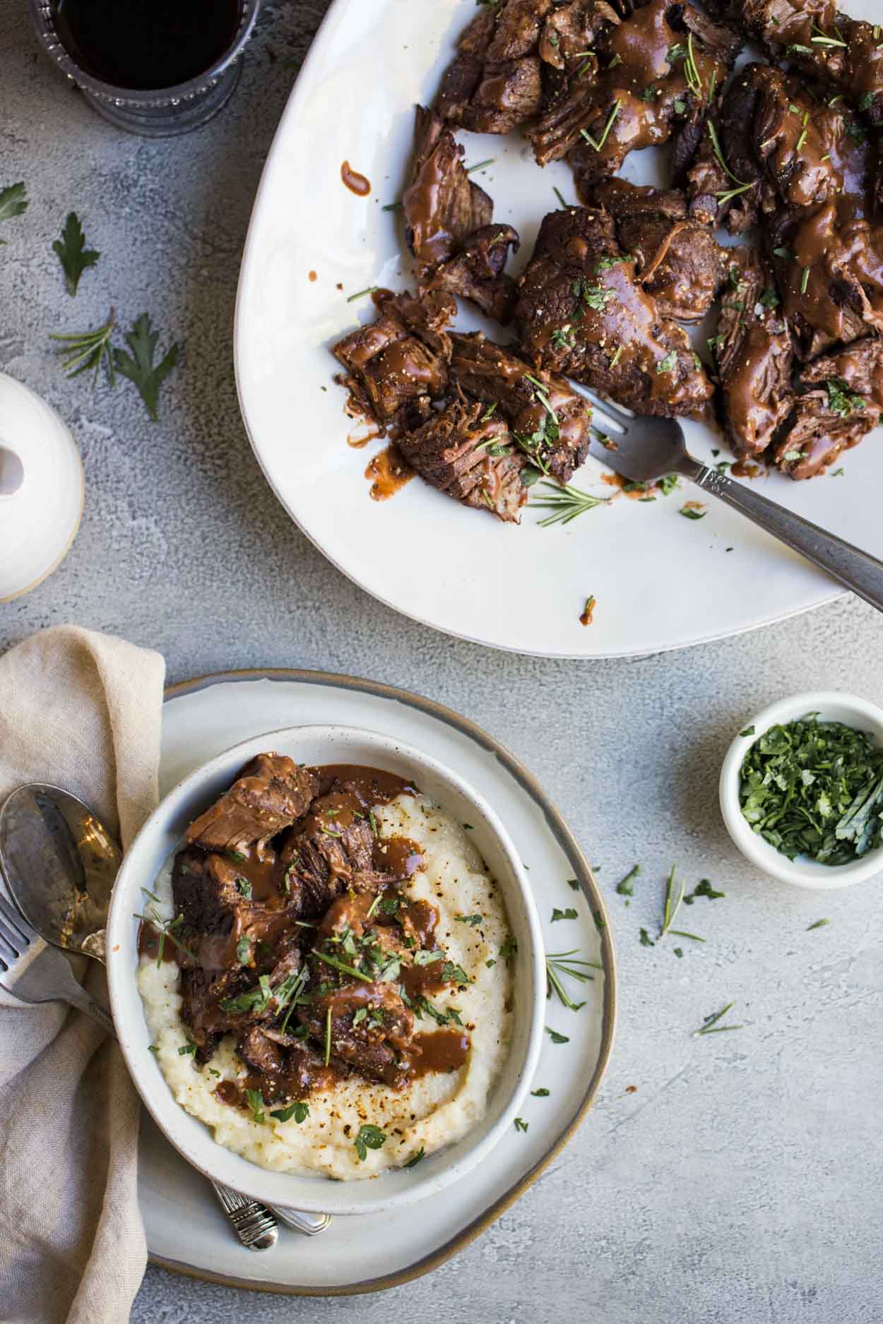 Pot roast and mashed potatoes with gravy in a white bowl and pot roast on a white platter with 2 wine glasses in the background