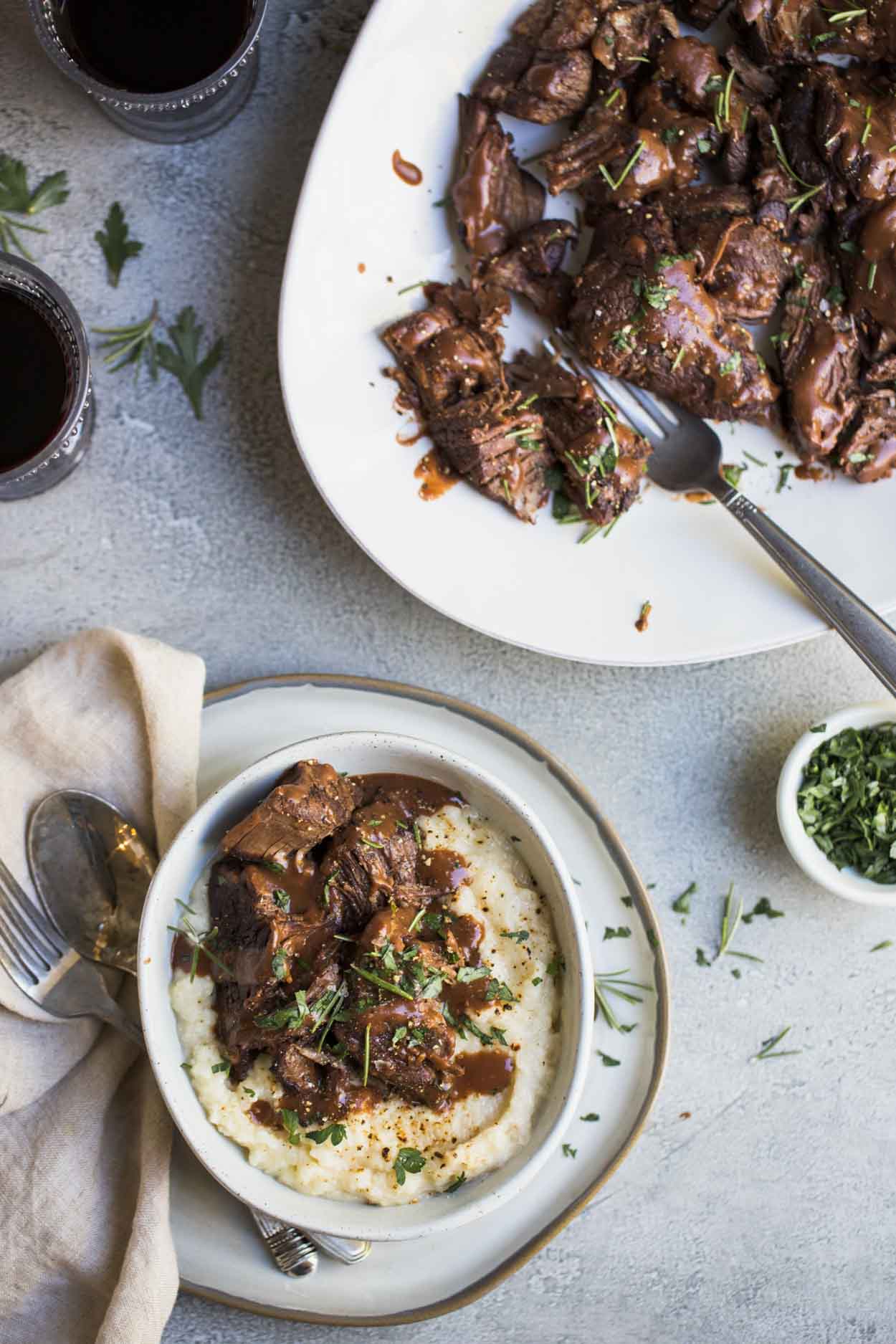 Pot roast and mashed potatoes with gravy in a white bowl and pot roast on a white platter with 2 wine glasses in the background 