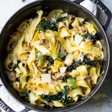 Wide egg noodles with sausage, kale and fennel in a black cast iron pot on a gray surface