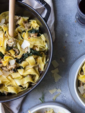 Wide egg noodles with sausage, kale and fennel in a black cast iron pot and on 2 gray bowls on a gray surface