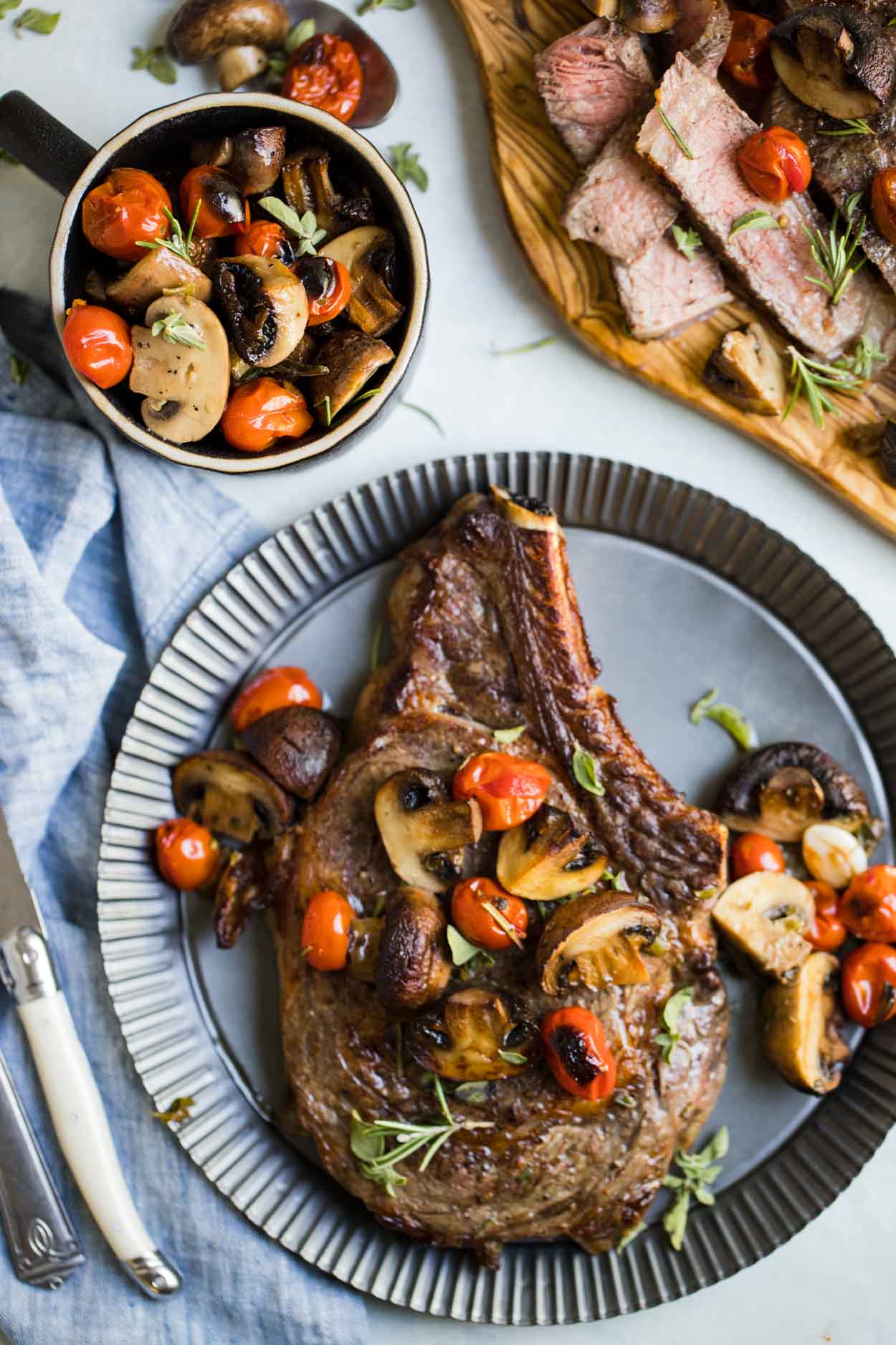 Steak, mushrooms, tomatoes and herbs on a tin plate with a bowl of mushrooms and tomatoes next to it and sliced meat on a cutting board, on top of a white background