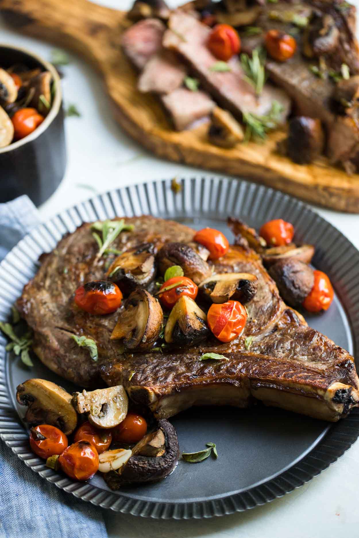 Steak, mushrooms, tomatoes and herbs on a tin plate with a bowl of mushrooms and tomatoes next to it and sliced meat on a cutting board, on top of a white background