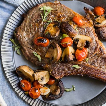 Steak, mushrooms, tomatoes and herbs on a tin plate with a bowl of mushrooms and tomatoes next to it and sliced meat on a cutting board, on top of a white background