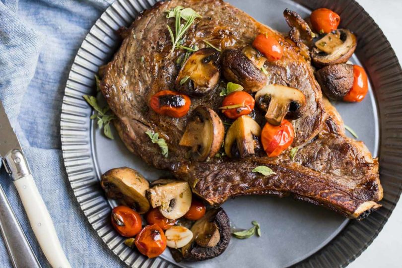 Steak, mushrooms, tomatoes and herbs on a tin plate with a bowl of mushrooms and tomatoes next to it and sliced meat on a cutting board, on top of a white background