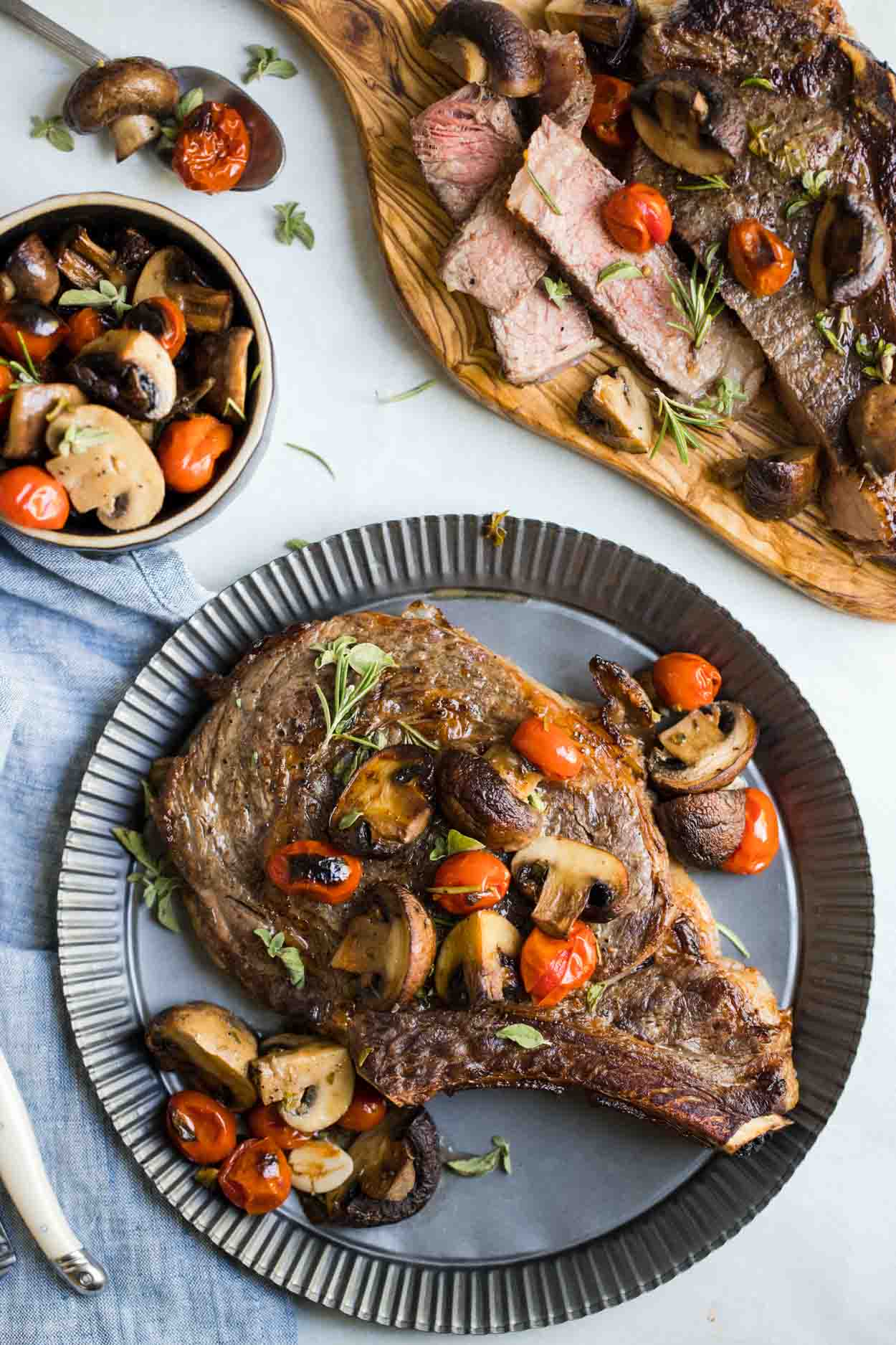 Steak, mushrooms, tomatoes and herbs on a tin plate with a bowl of mushrooms and tomatoes next to it and sliced meat on a cutting board, on top of a white background 