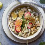 beige bowl filled with pork posole soup and garnished with limes, cabbage and radishes, with a silver spoon set to the side