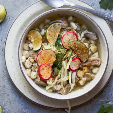 beige bowl filled with pork posole soup and garnished with limes, cabbage and radishes, with a silver spoon set to the side