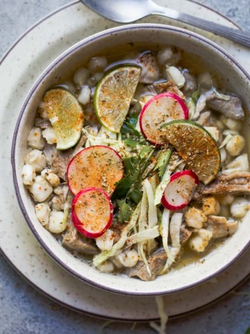 beige bowl filled with pork posole soup and garnished with limes, cabbage and radishes, with a silver spoon set to the side