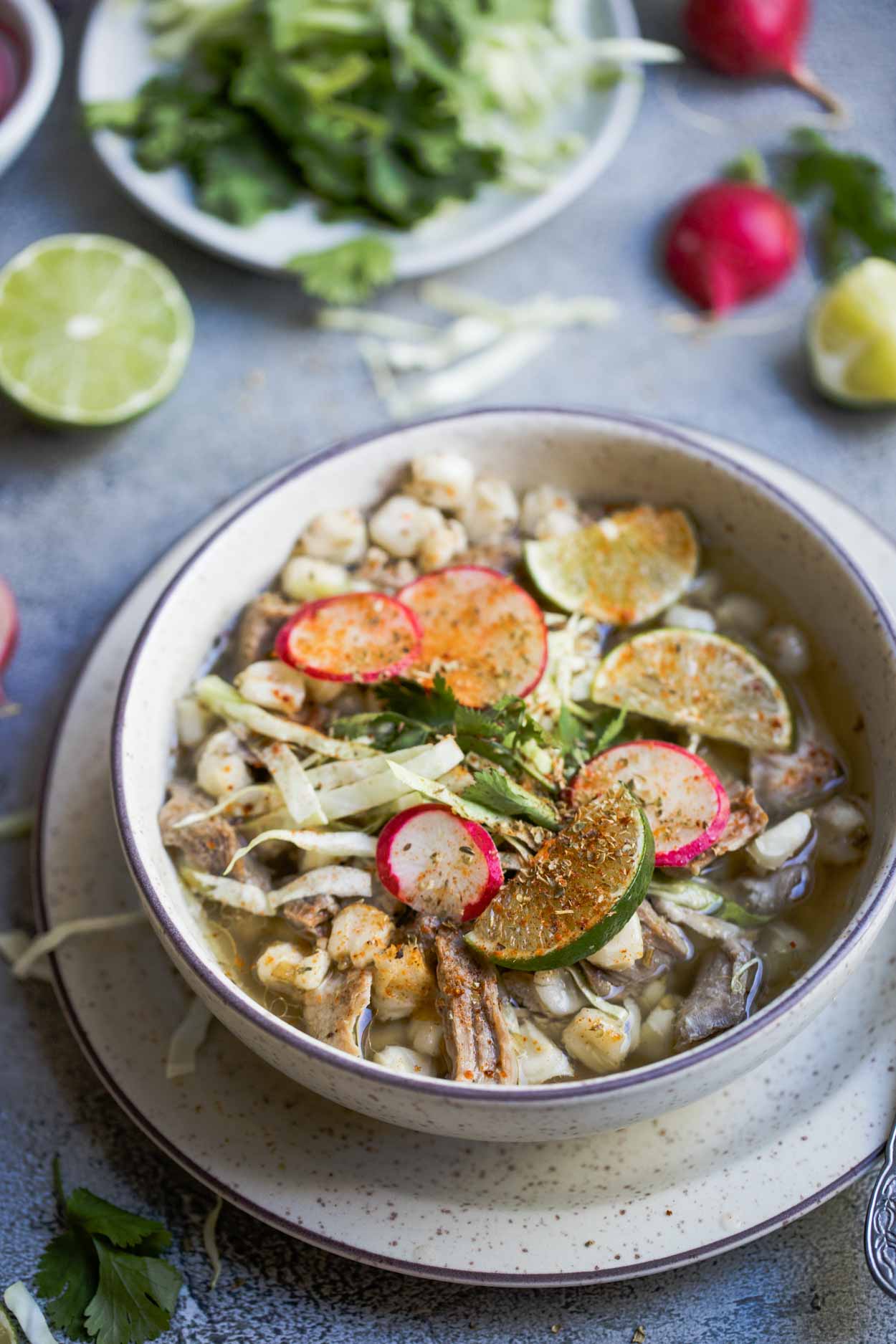 Beige bowl filled with posole soup and various garnishes, set on a beige plate, against a gray background