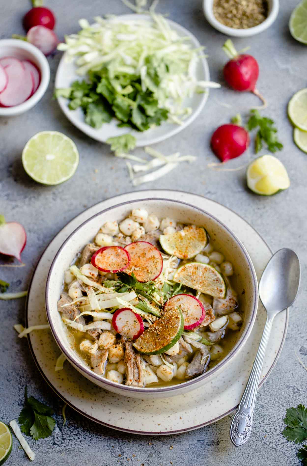 Warm posole soup filled in beige bowl, set on a gray table with lime, cabbage and radishes scattered about