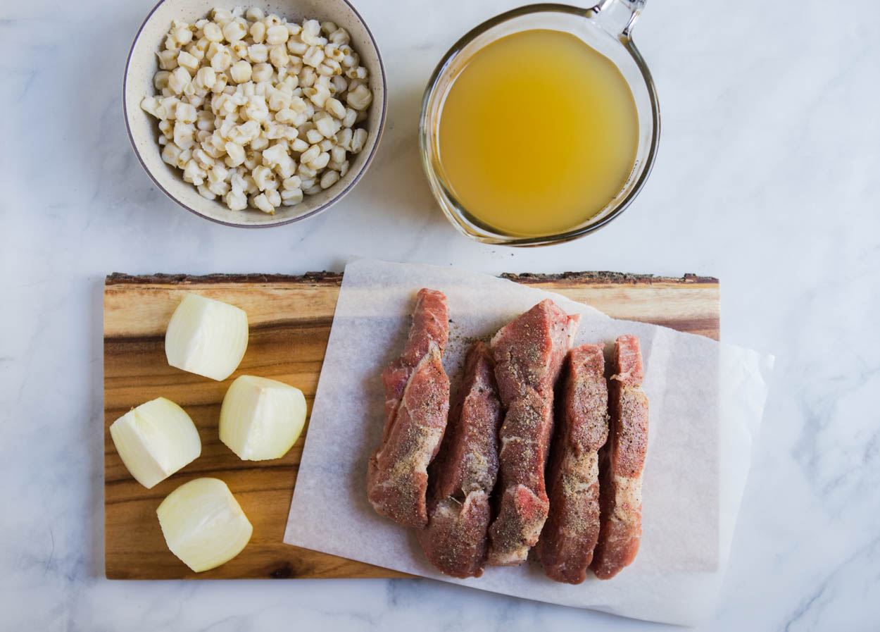 pieces of country style pork ribs set on a piece of parchment paper, alongside cut up onion, a bowl of hominy, and chicken broth