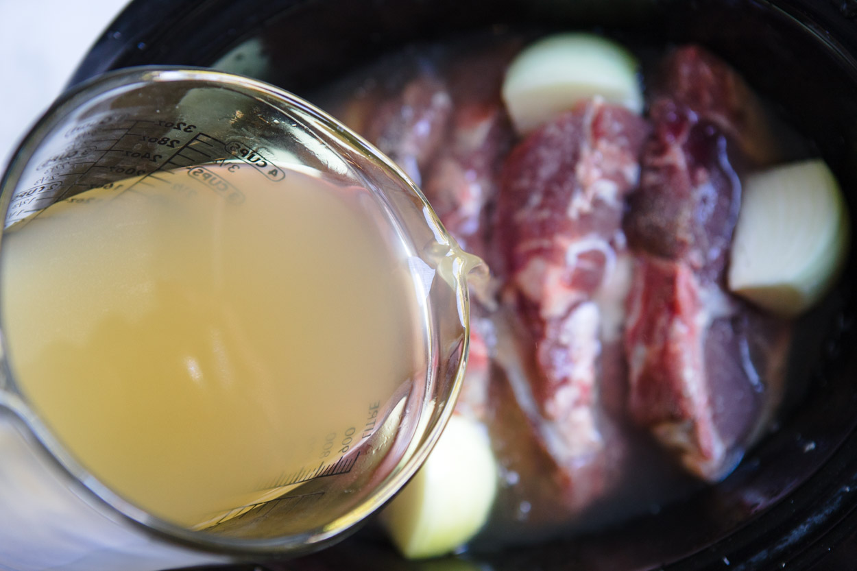 chicken broth being poured over pork ribs in a slow cooker