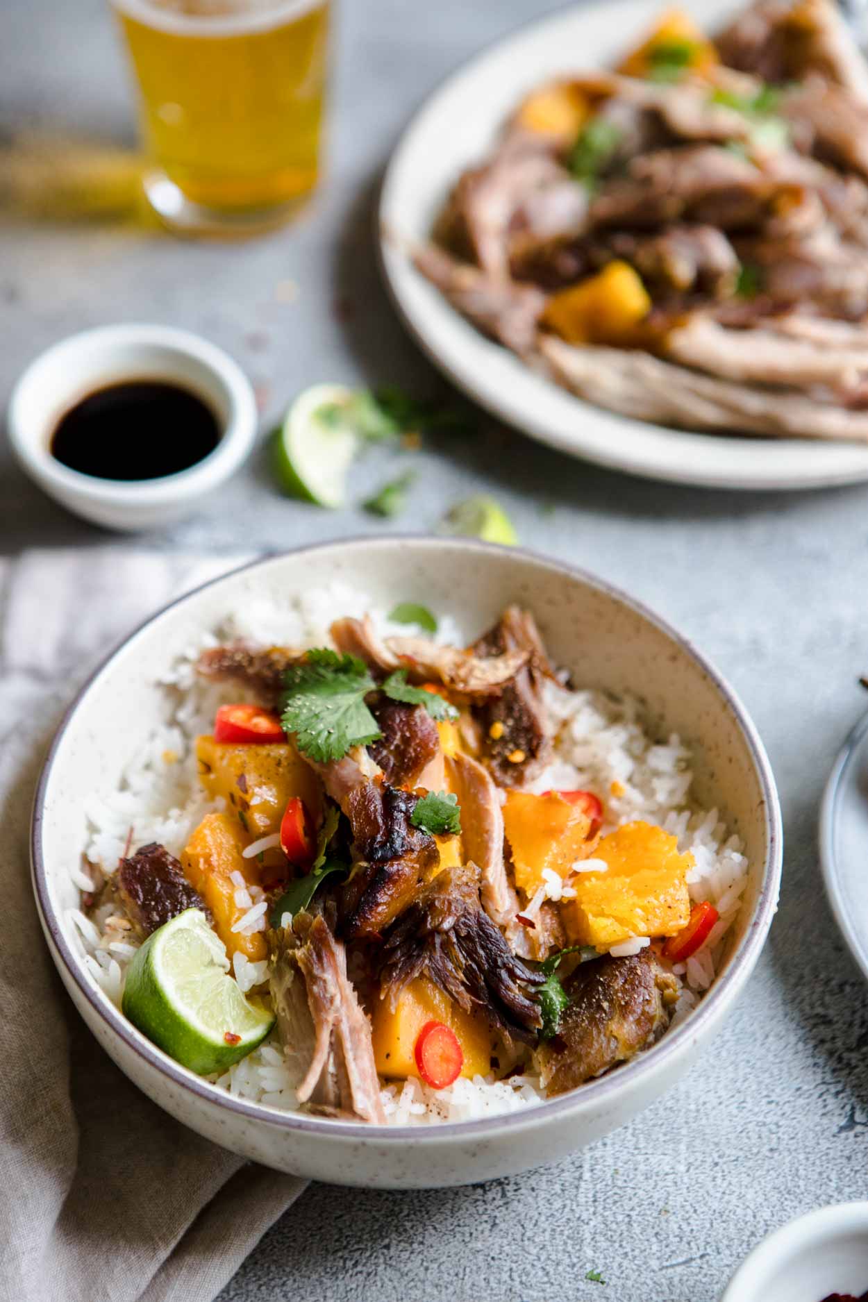 platter of braised Asian pork shoulder sitting next to a bowl filled with rice and pork shoulder and a glass of beer