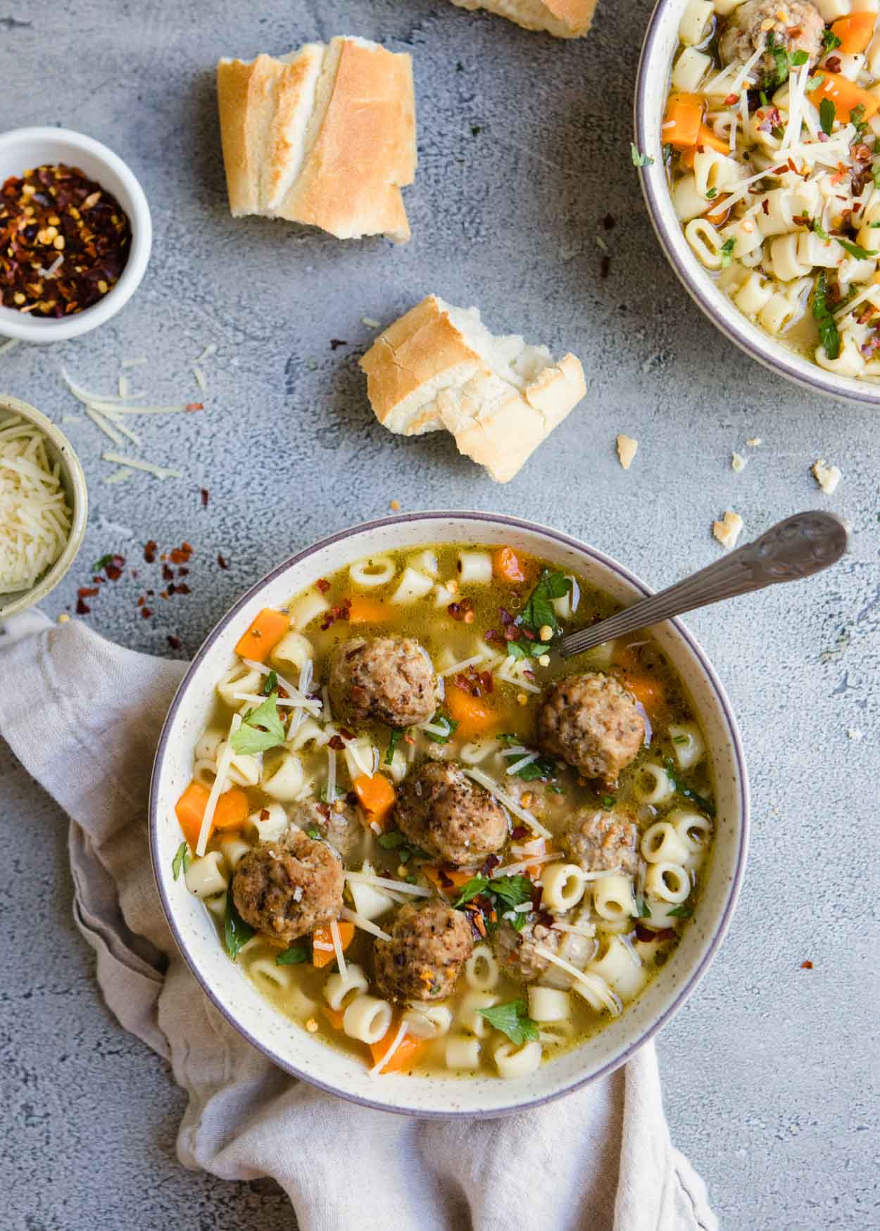 Round bowl filled with pasta and meatball soup and chili flakes, Parmesan cheese and crusty bread