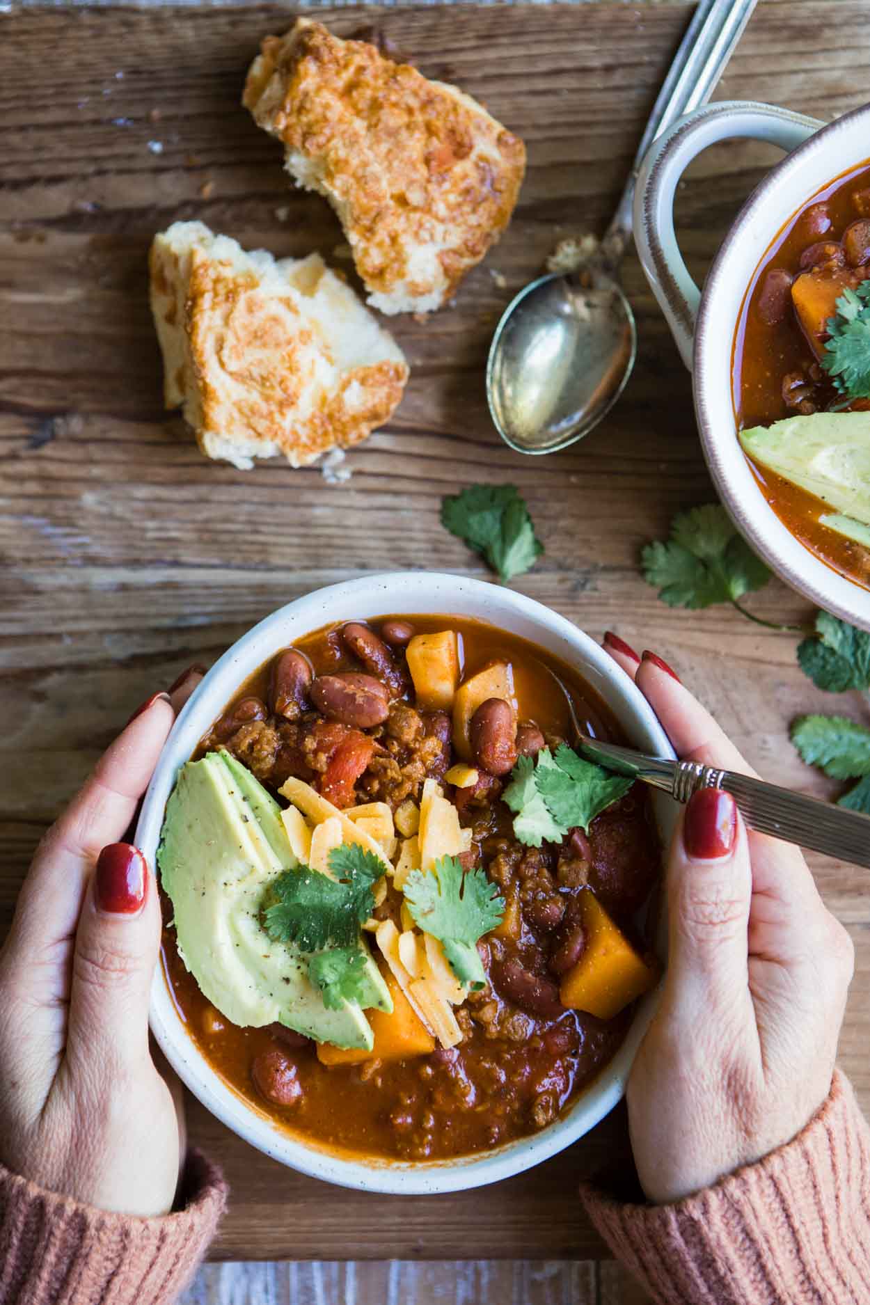 a woman's hand cradling a warm bowl of sweet potato vegetarian chili garnished with avocado, cheddar cheese and cilantro