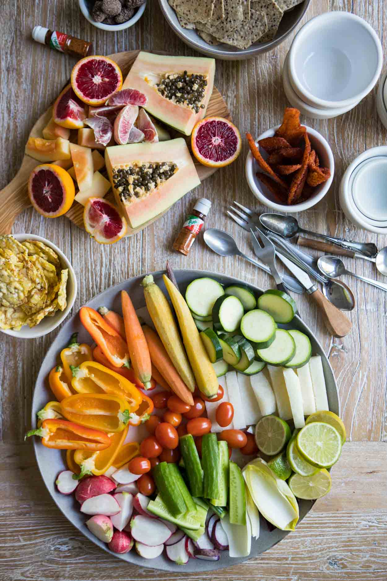 A mix of fruits and veggies on a round platter along with some other utensils and snacks to make a crudite