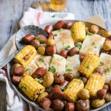 round platter with sausage, halibut, corn and potatoes set against a gray backdrop