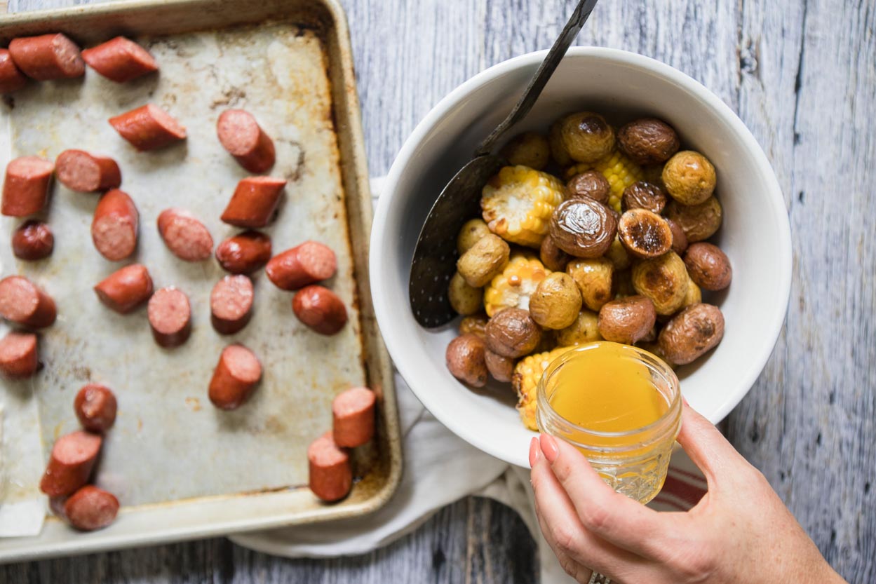 Butter sauce being poured over potatoes and corn next to a tray of sausage 