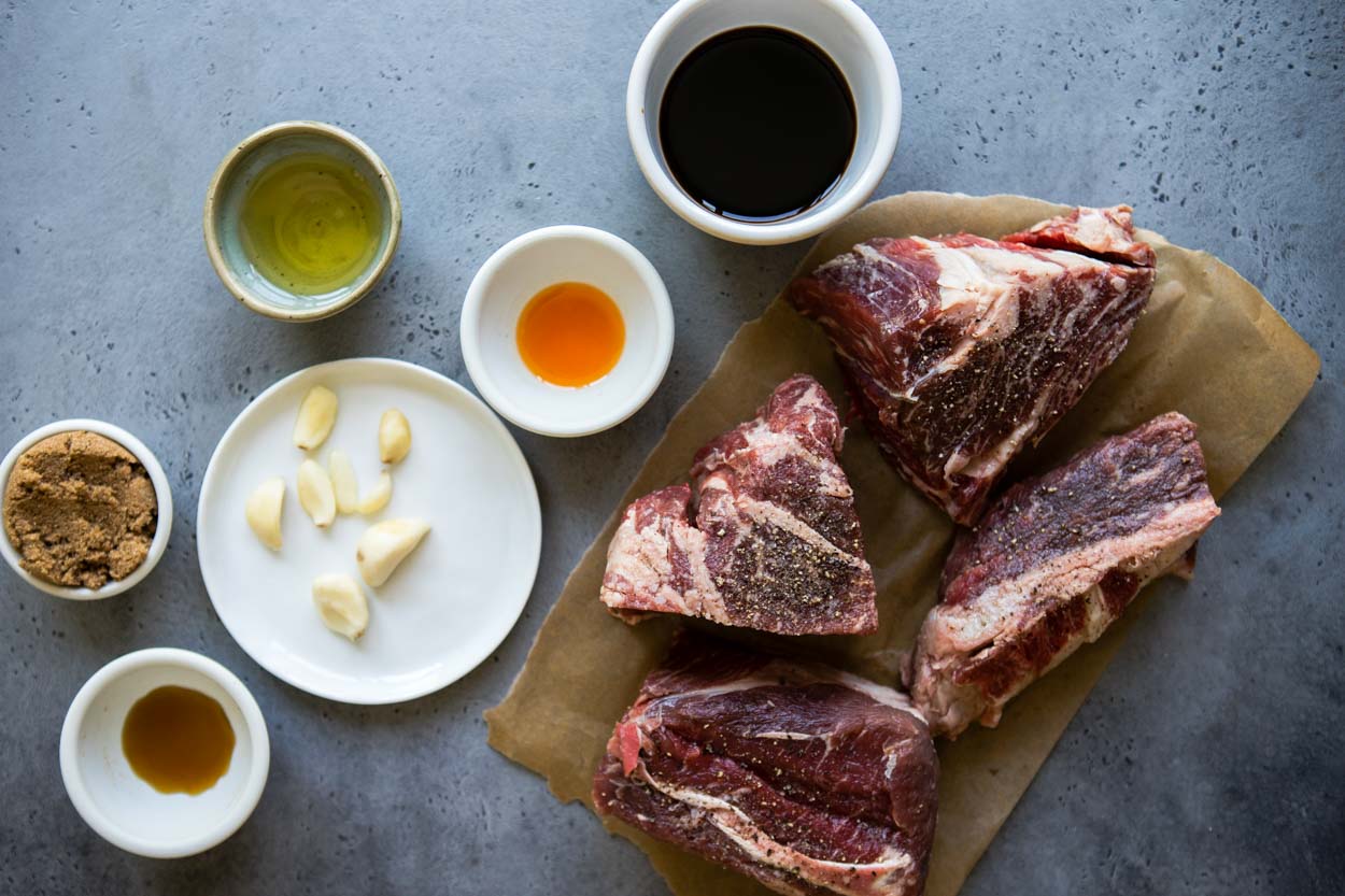 soy sauce, sesame oil, fish sauce, brown sugar and chili oil in little bowls next to a piece of chuck roast and a plate of garlic