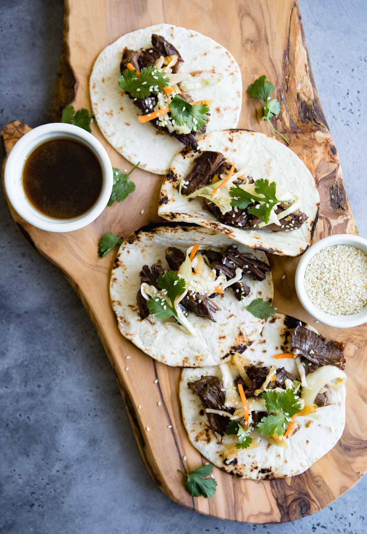 Korean Beef Tacos on 4 flour tortillas and sauce on a wooden board set against a gray background