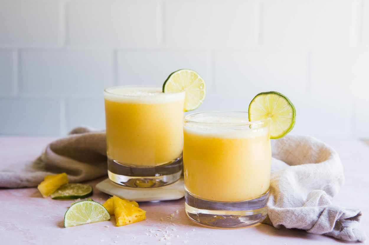 2 glasses filled with pineapple margarita set on a pink table with lime and pineapple slices as garnish