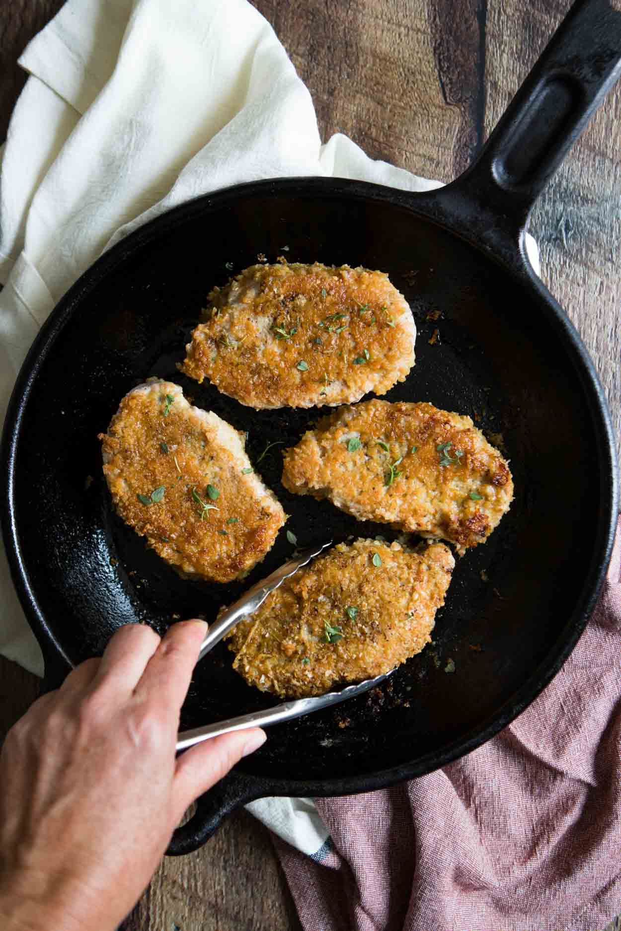 four parmesan pork chops in a cast iron skillet and a hand turning one over with tongs