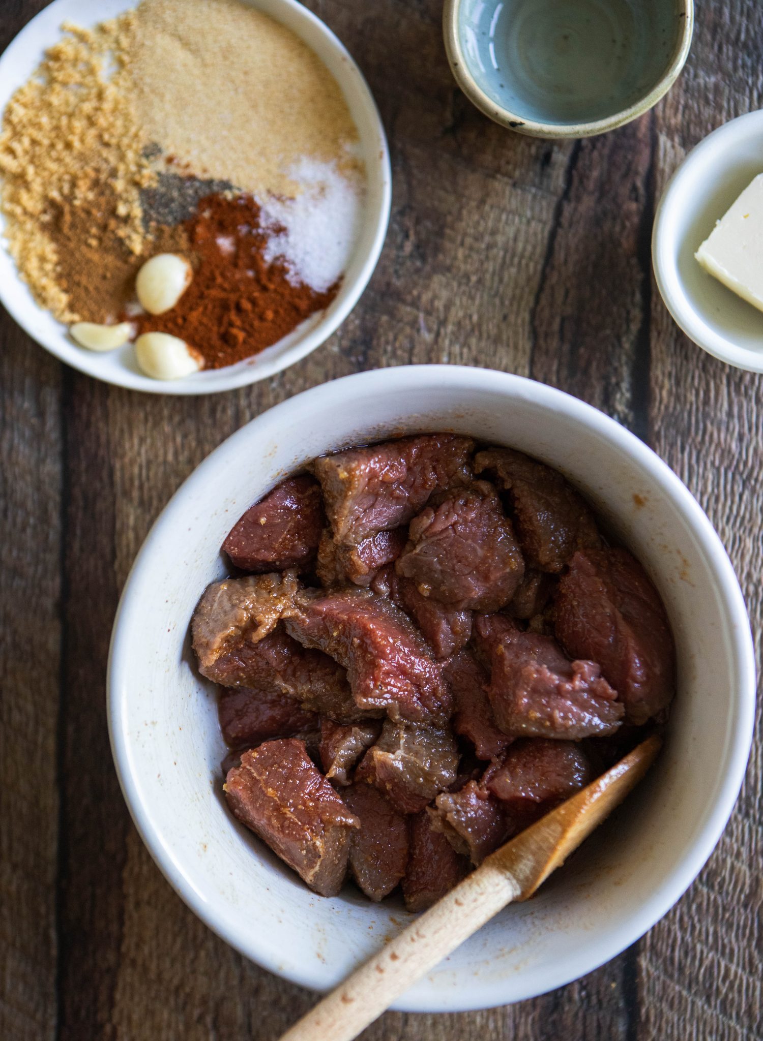 bowl of beef sirloin steak tips mixed with various herbs and spices, set next to a plate of herbs and spices, on a wooden background