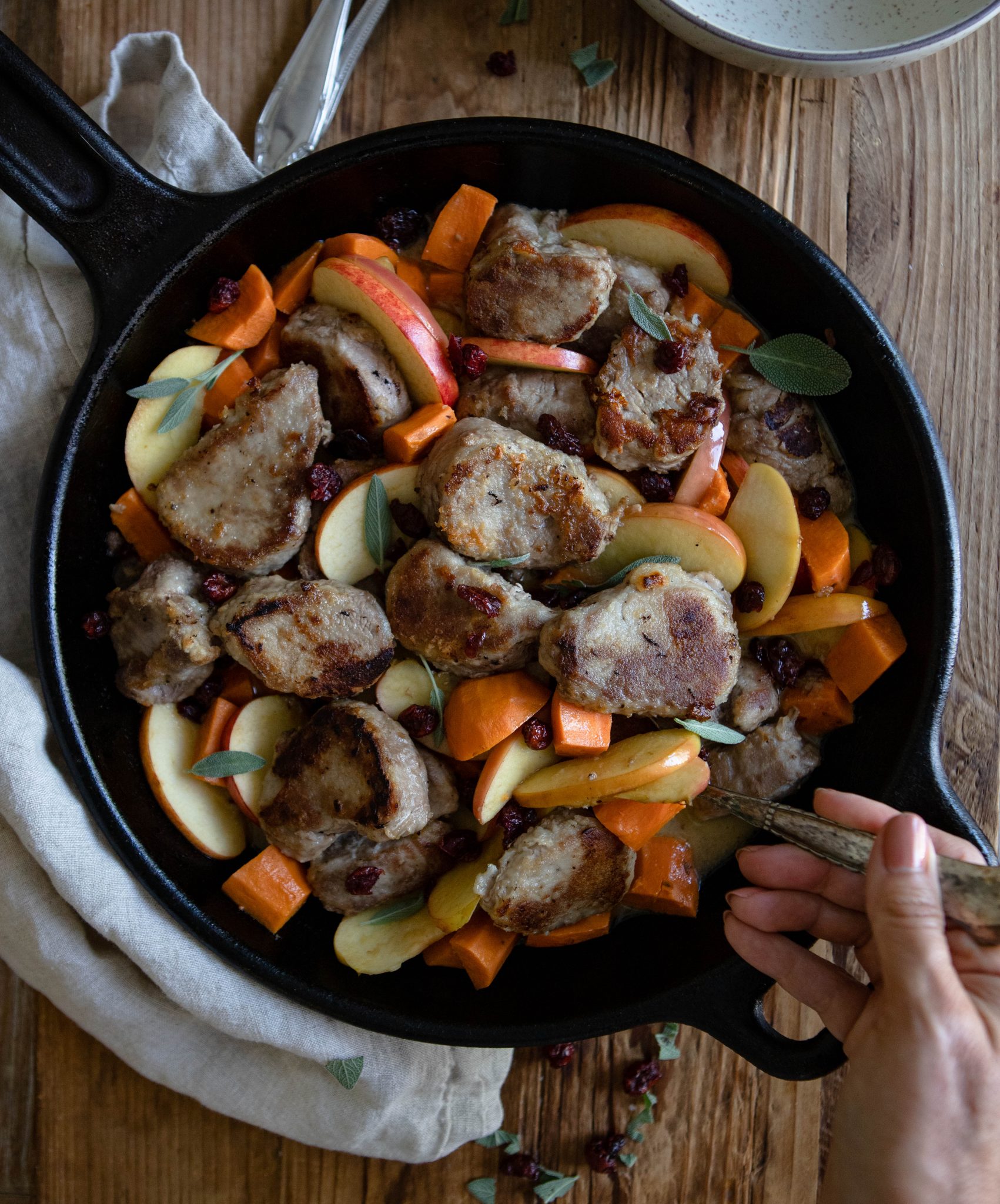 Woman's hand holding a spoon to serve a pork tenderloin dish out of a cast iron skillet