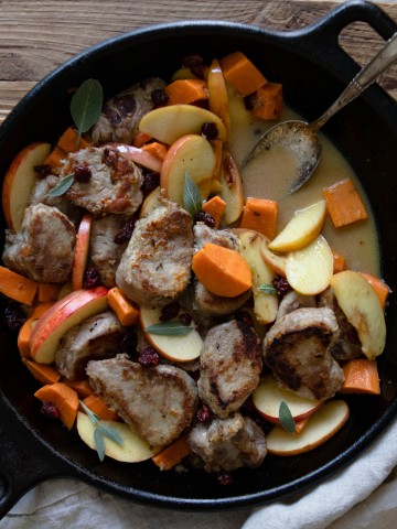 black cast iron skillet with pork tenderloin, apples and sweet potatoes on a wooden board