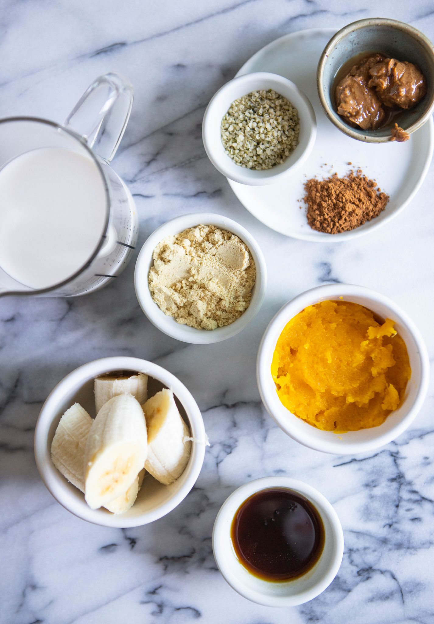 ingredients for pumpkin spice smoothie in little white bowls and glass jars, set against a marble backdrop