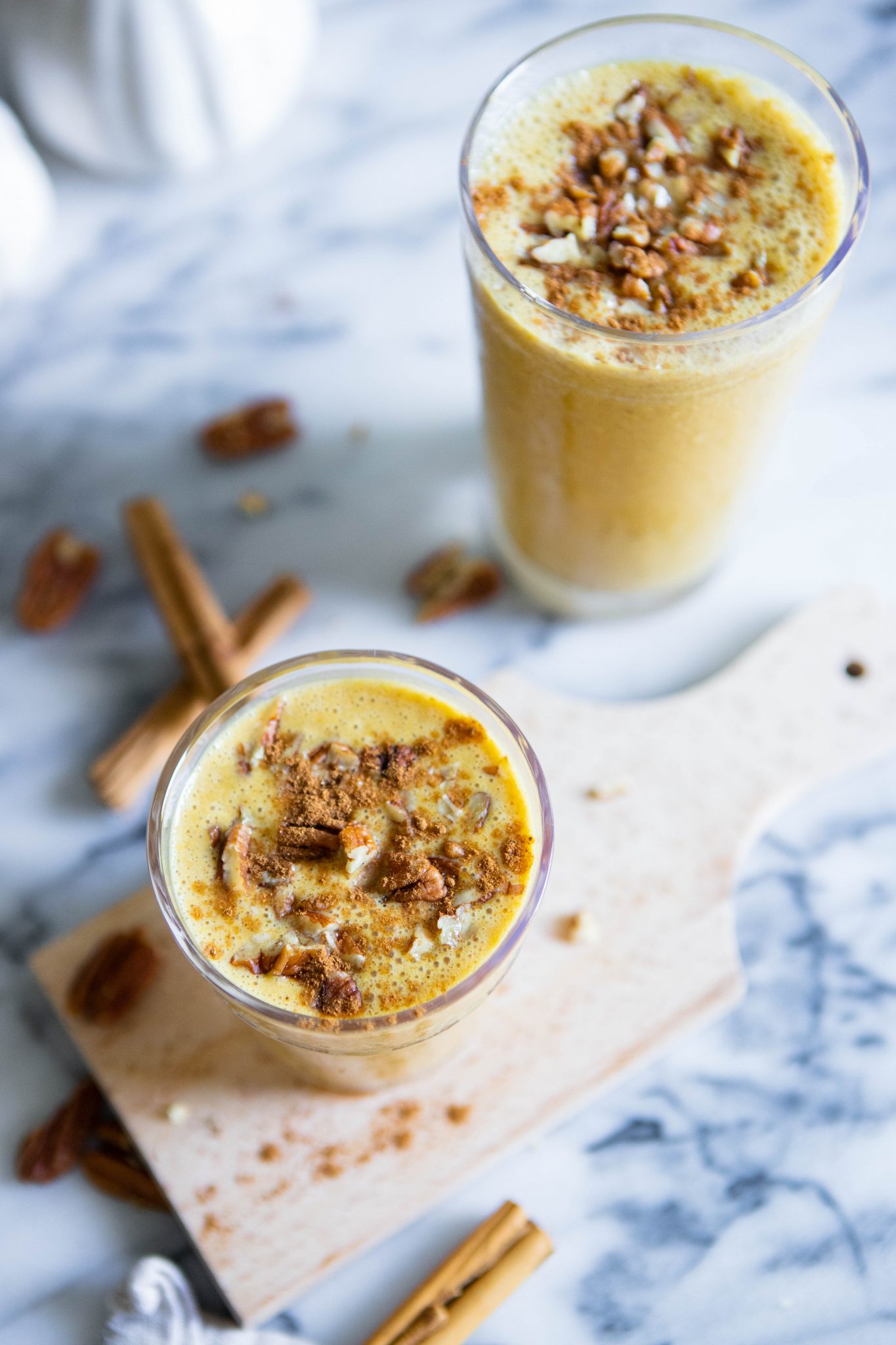 2 glasses filled with pumpkin smoothie set on a marble backdrop with pecans and cinnamon sticks