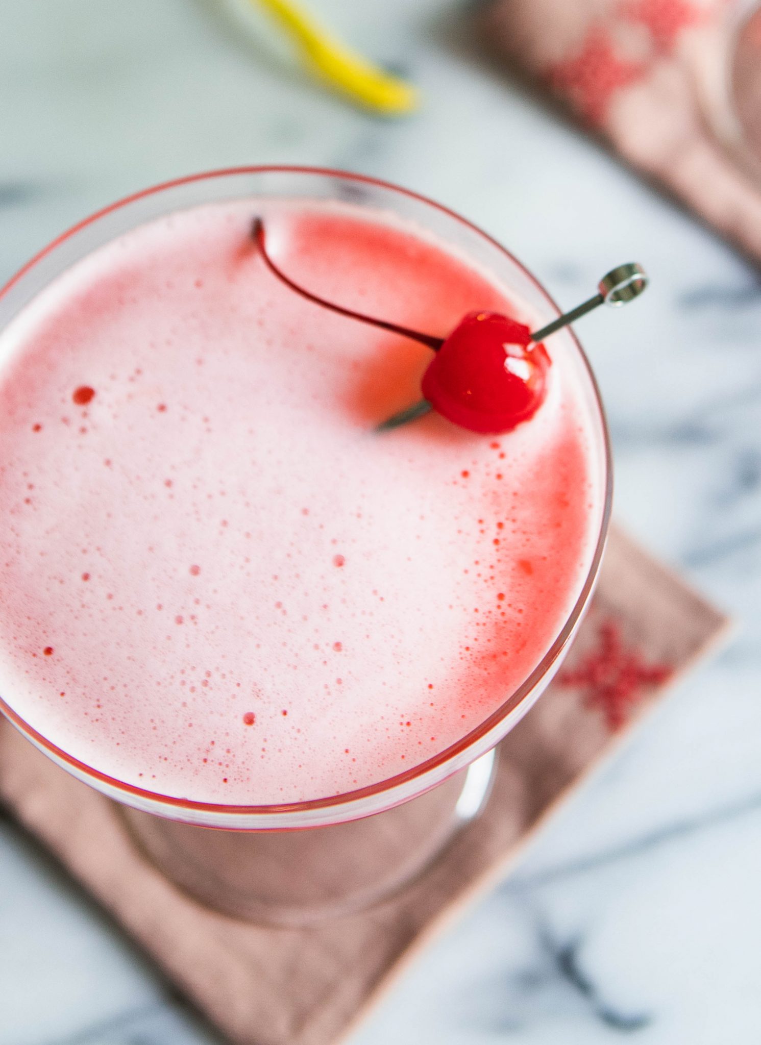 pink foamy cocktail in a coupe glass garnished with maraschino cherry