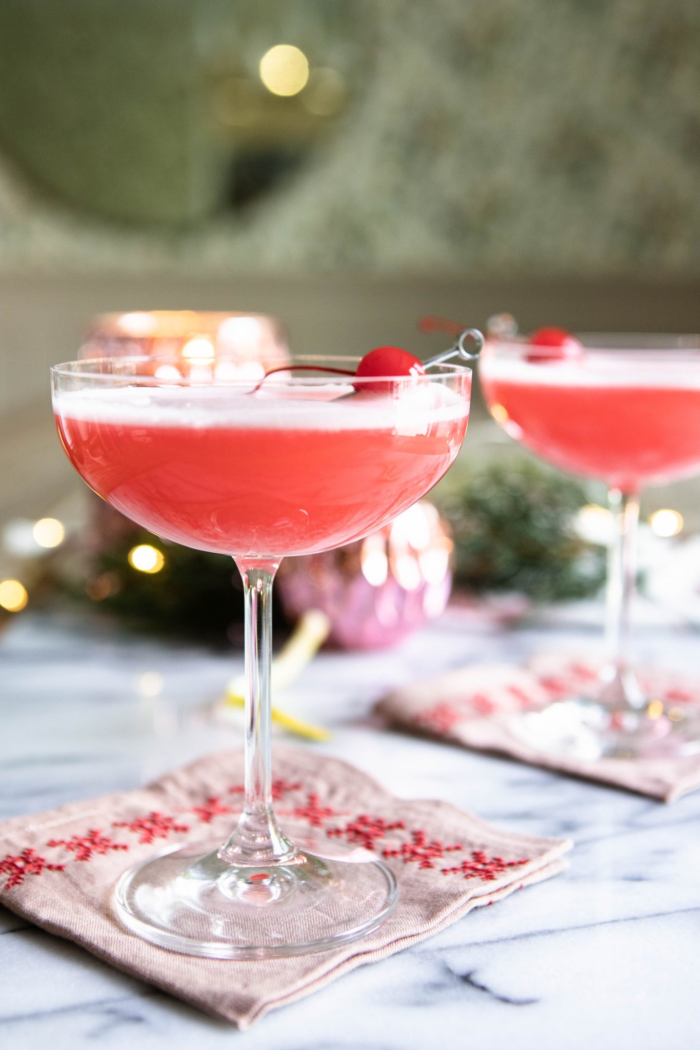 2 coupe glasses filled with a pink cocktail and garnished with a maraschino cherry and lemon peel, against a marble backdrop
