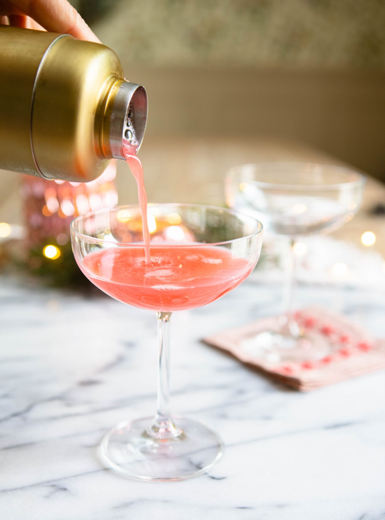 pink cocktail being poured into a coupe glass from a gold shaker