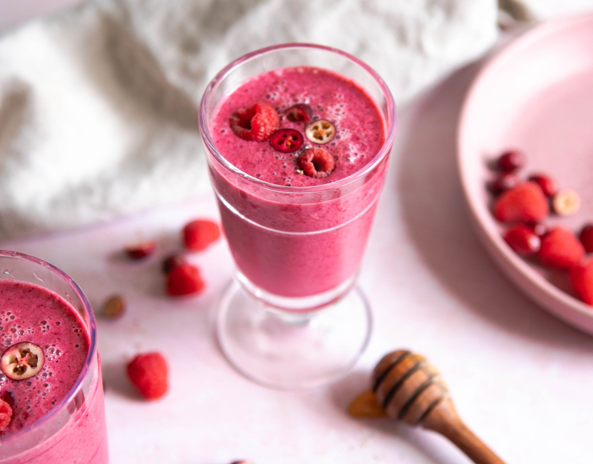 bright pink smoothie in a glass surrounded by honey, berries against a pink background