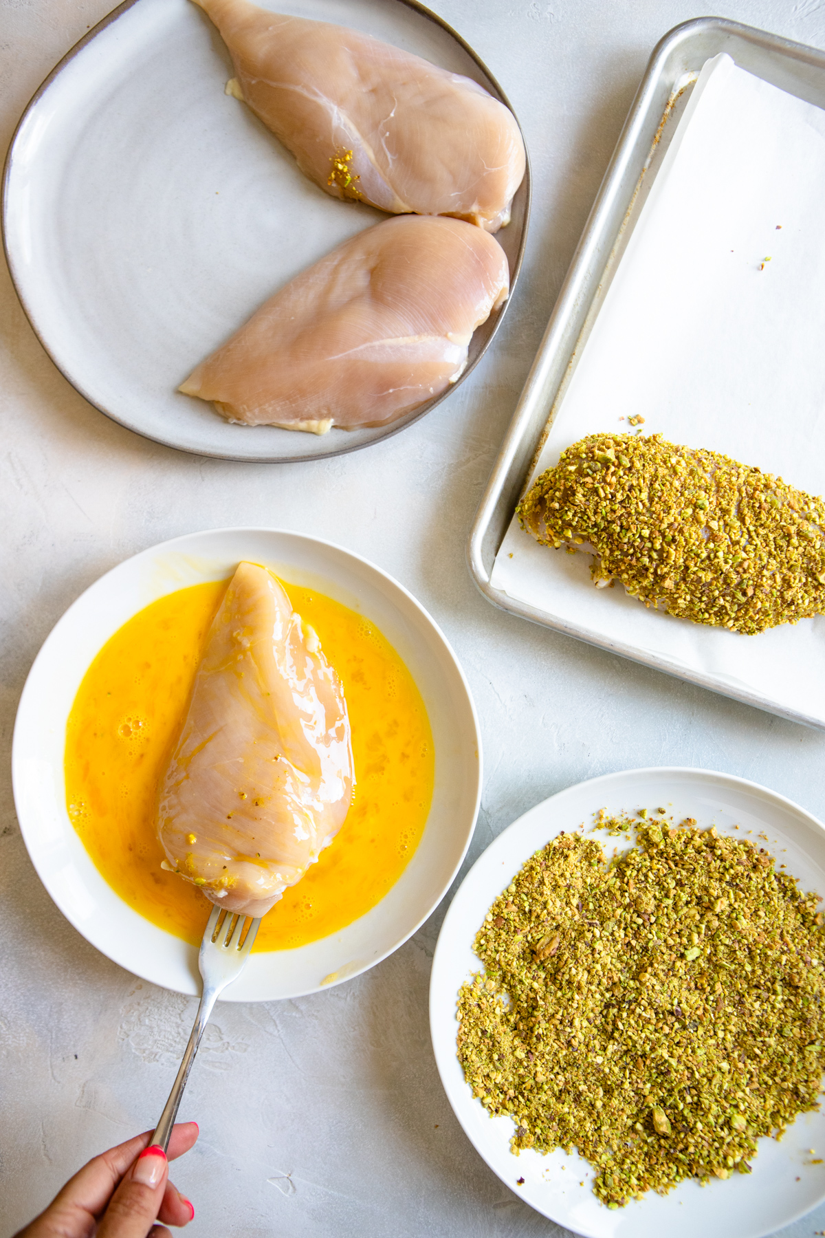 egg coating in a bowl next to a shallow dish of pistachios and chicken breasts being coated in egg