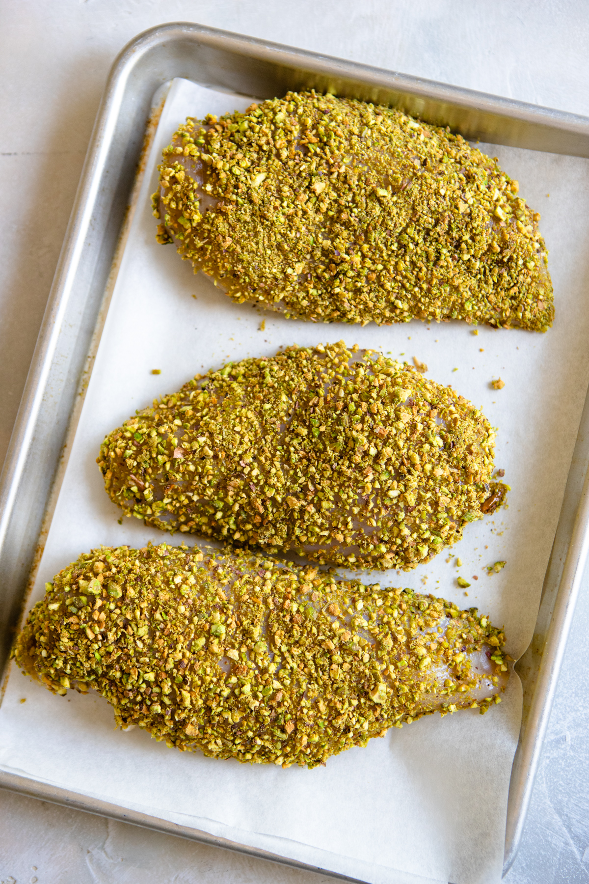 3 chicken breasts coated in pistachios on a parchment lined baking sheet