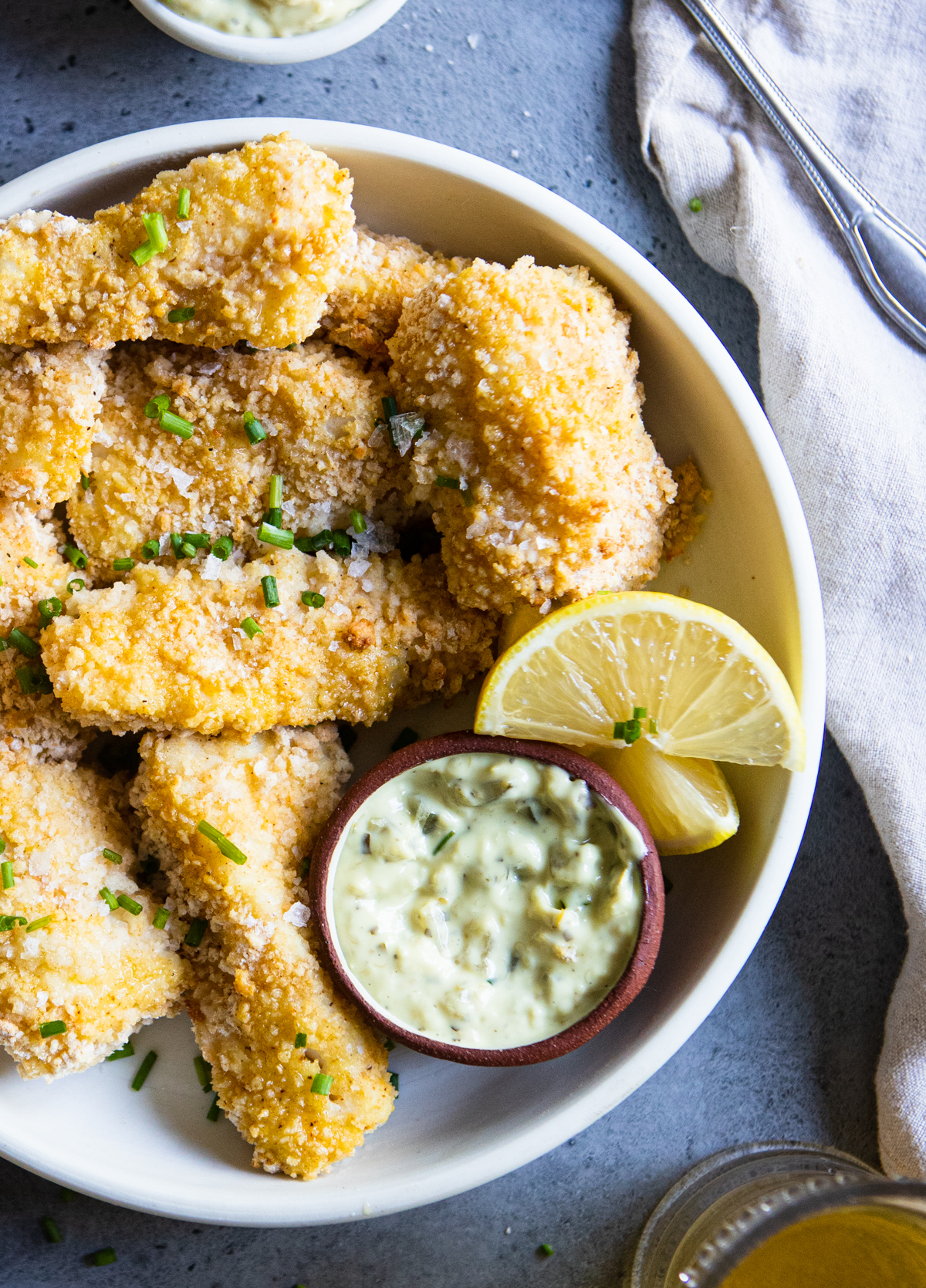 breaded cod set on a white plate against a gray back drop and garnished with lemon wedges and tartar sauce