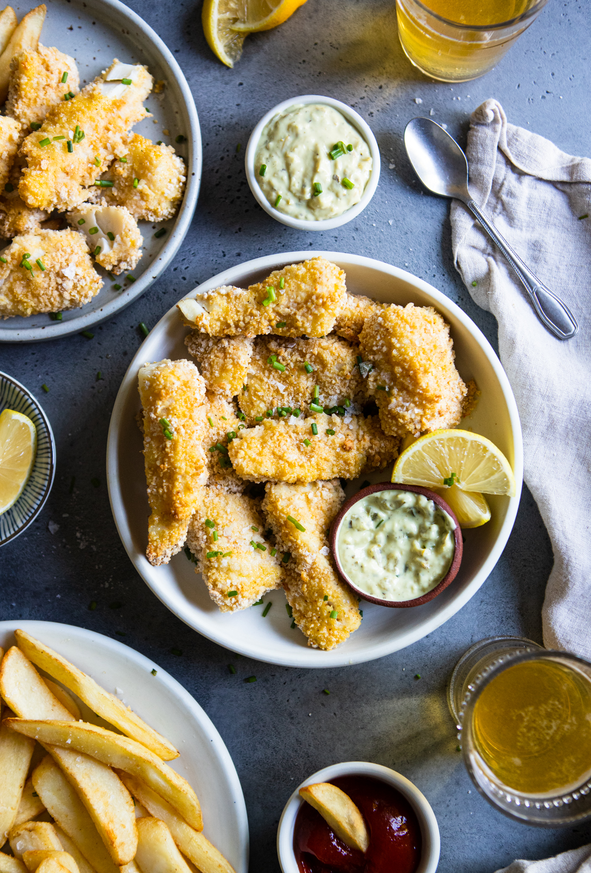 round white plate filled with fish sticks in air fryer pieces next to a bowl of tartar sauce and glasses of beer