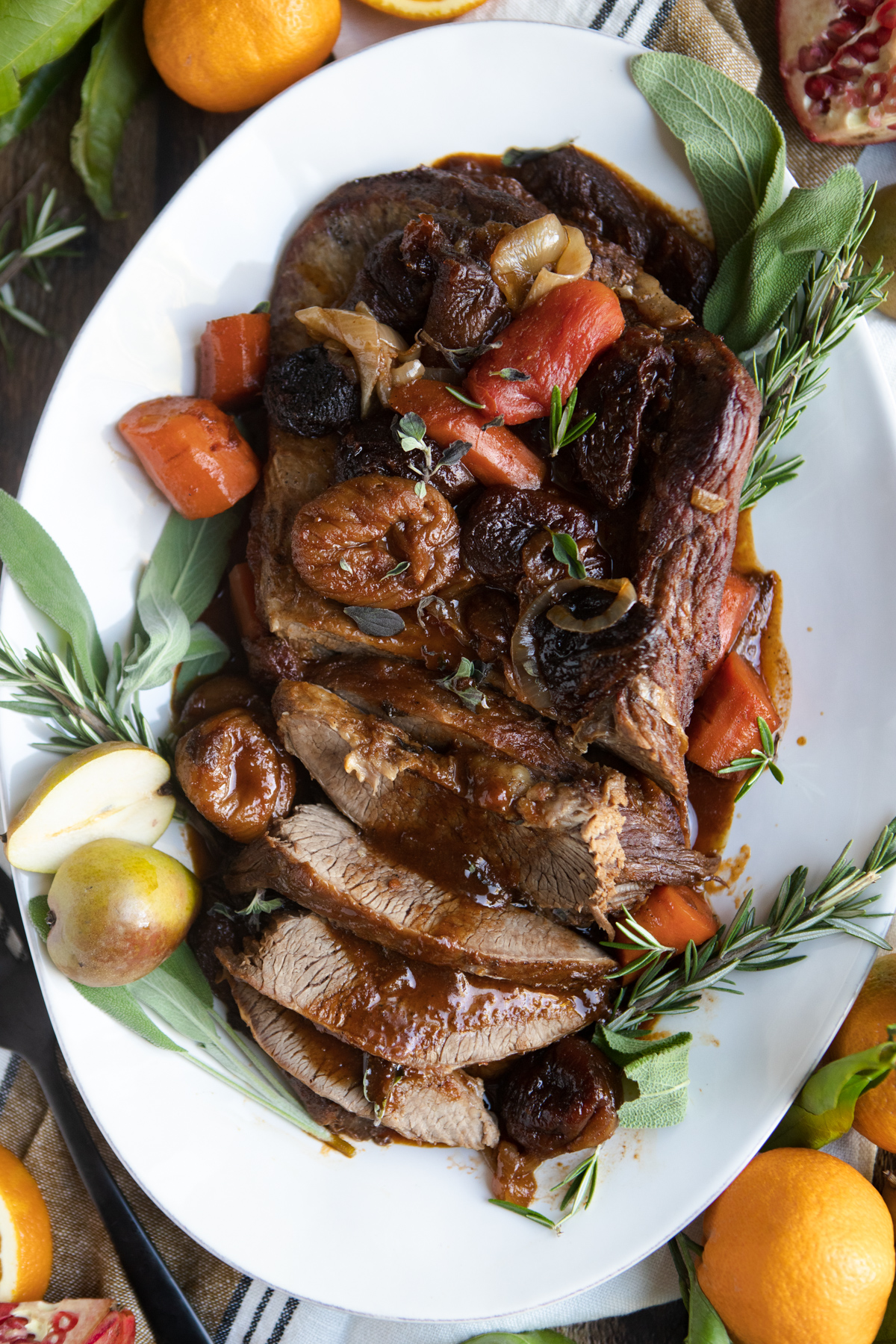 Christmas brisket recipe on a white platter surrounded by rosemary garnishes and topped with rich wine sauce and dried fruit 