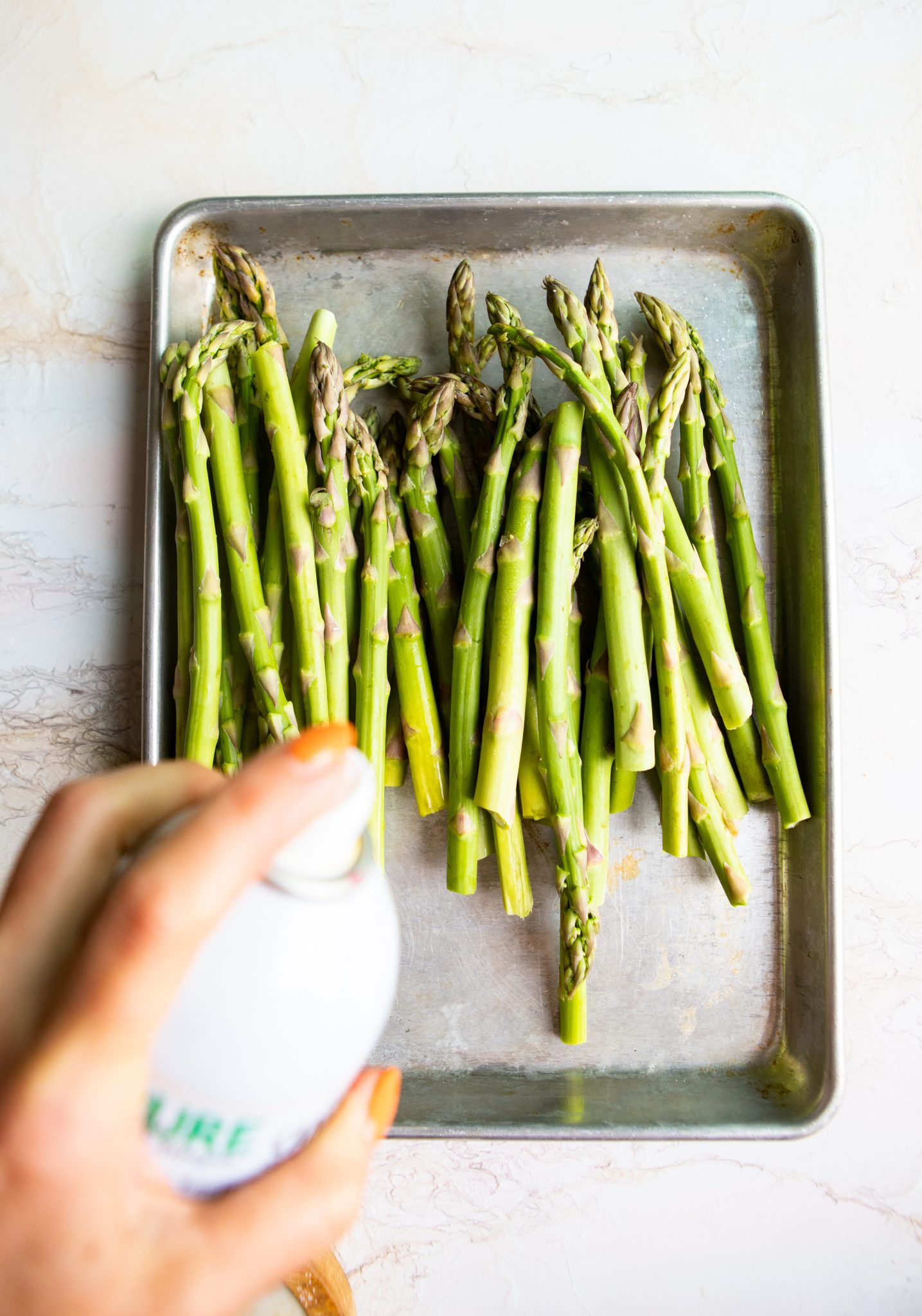 raw asparagus spears on a baking sheet with cooking spray being sprayed on them