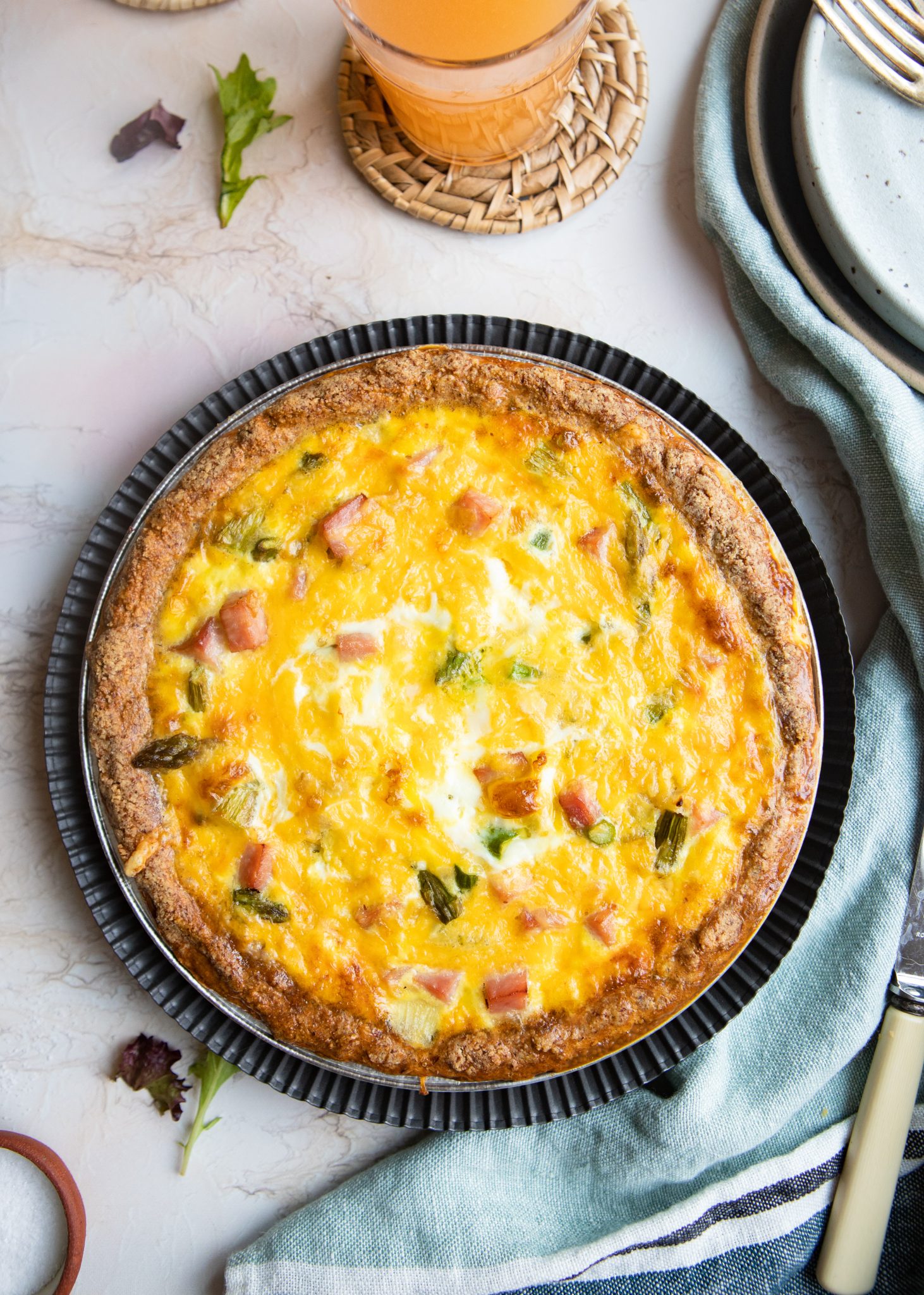 baked low carb ham and asparagus quiche on a metal pie plate and next to a stack of plates and grapefruit juice