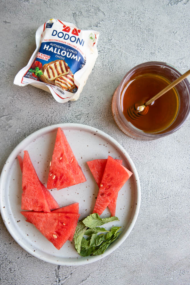 block of halloumi cheese next to a plate of sliced watermelon and mint leaves with a jar of honey