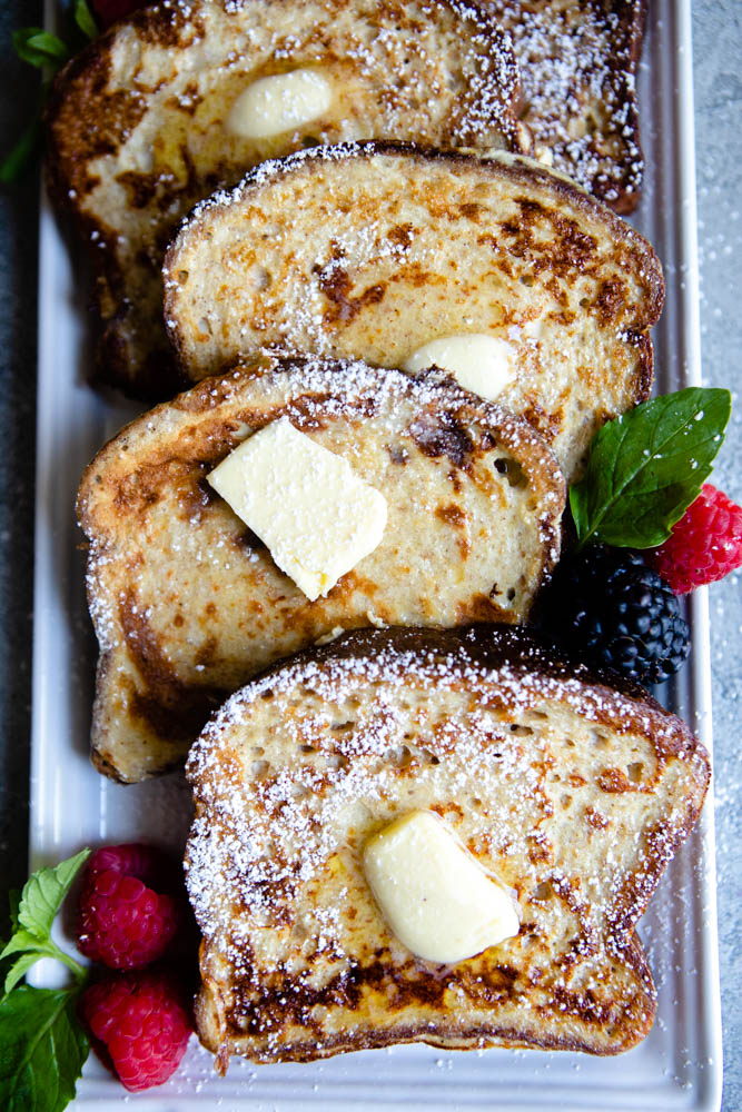 thick slices of high protein french toast layered on a white plate topped with butter and powdered sugar